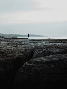 A lone figure stands on a rocky coastline under an overcast sky, creating a sense of isolation and tranquility.