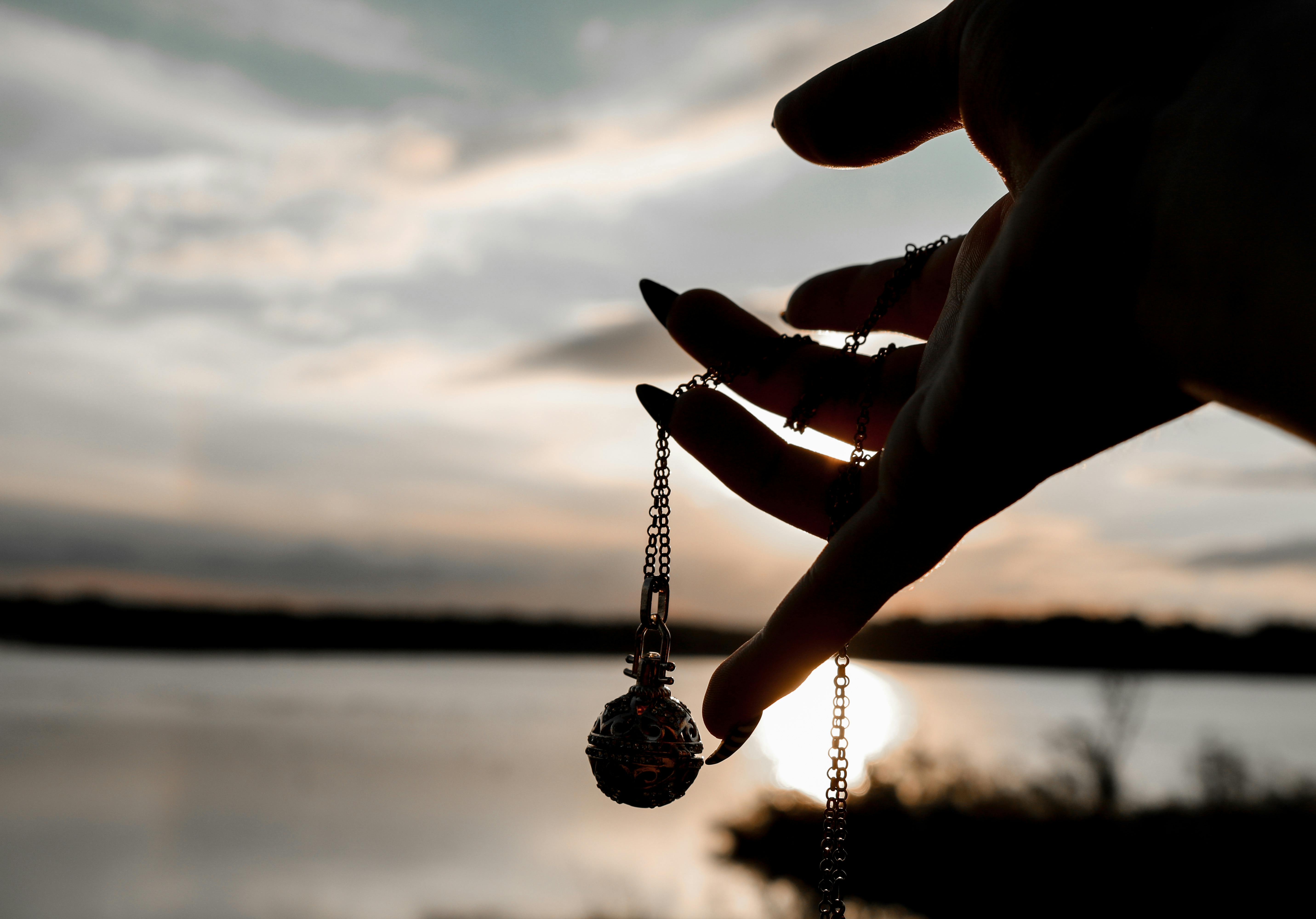 Silhouette of a hand holding a pendant necklace by the water during a serene sunset.