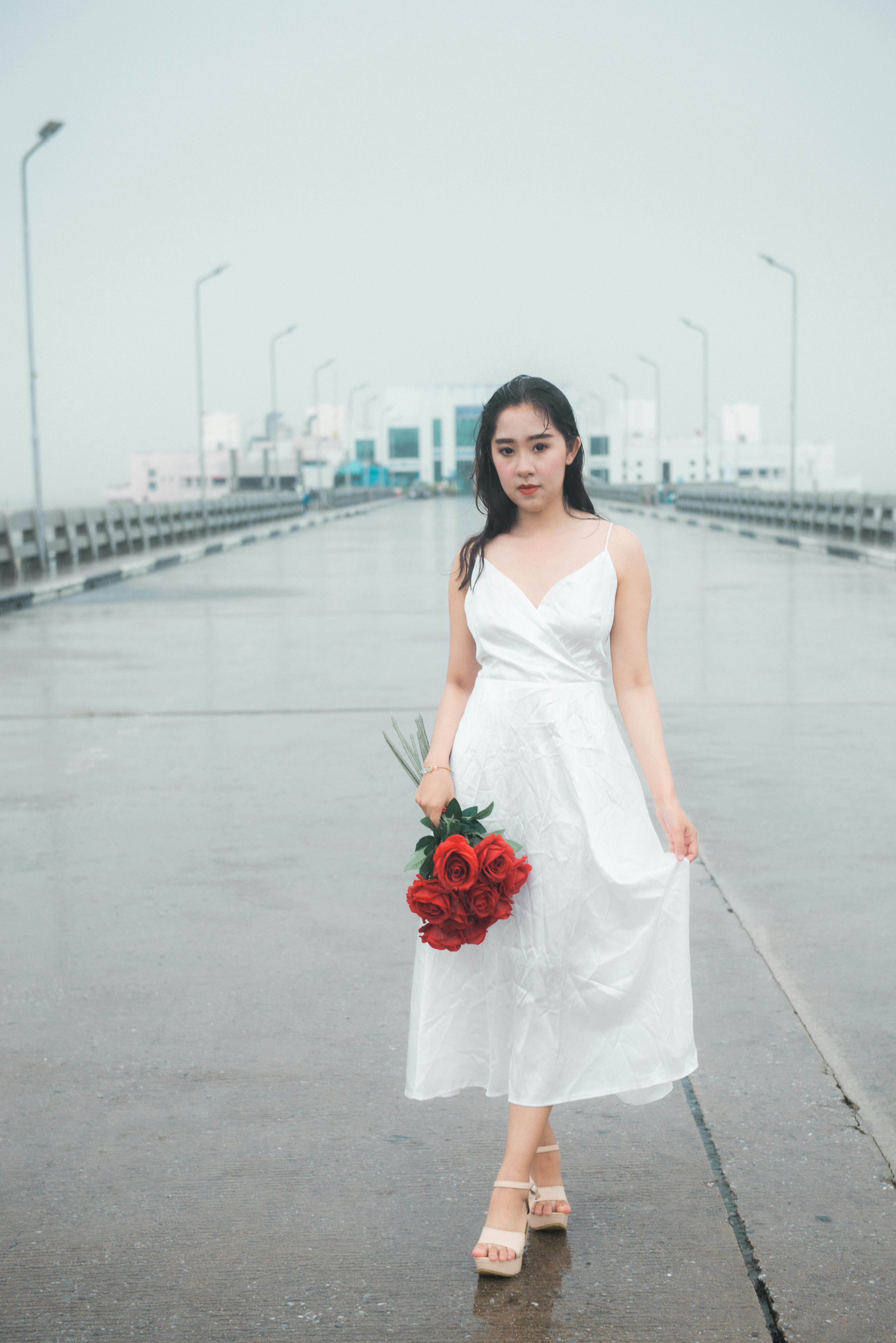 A woman in a white dress holding red roses stands on a bridge, with a serene background.