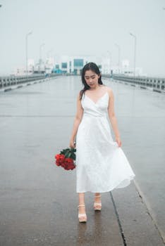 Asian woman in a white dress holding red roses on a rainy day, walking on a wet pier.