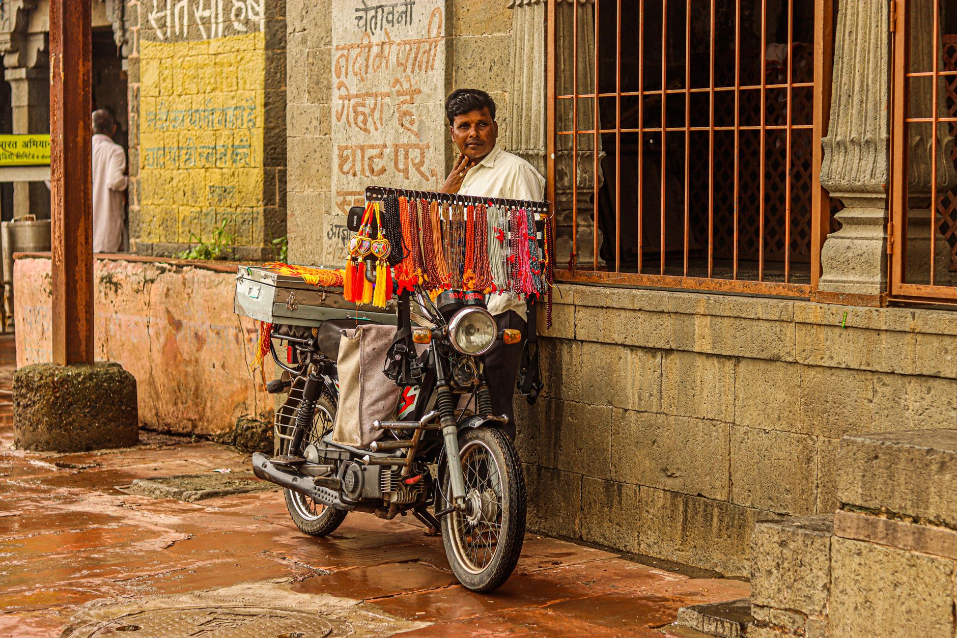 A man sells vibrant necklaces from his motorcycle on a wet street, showcasing local craftsmanship.