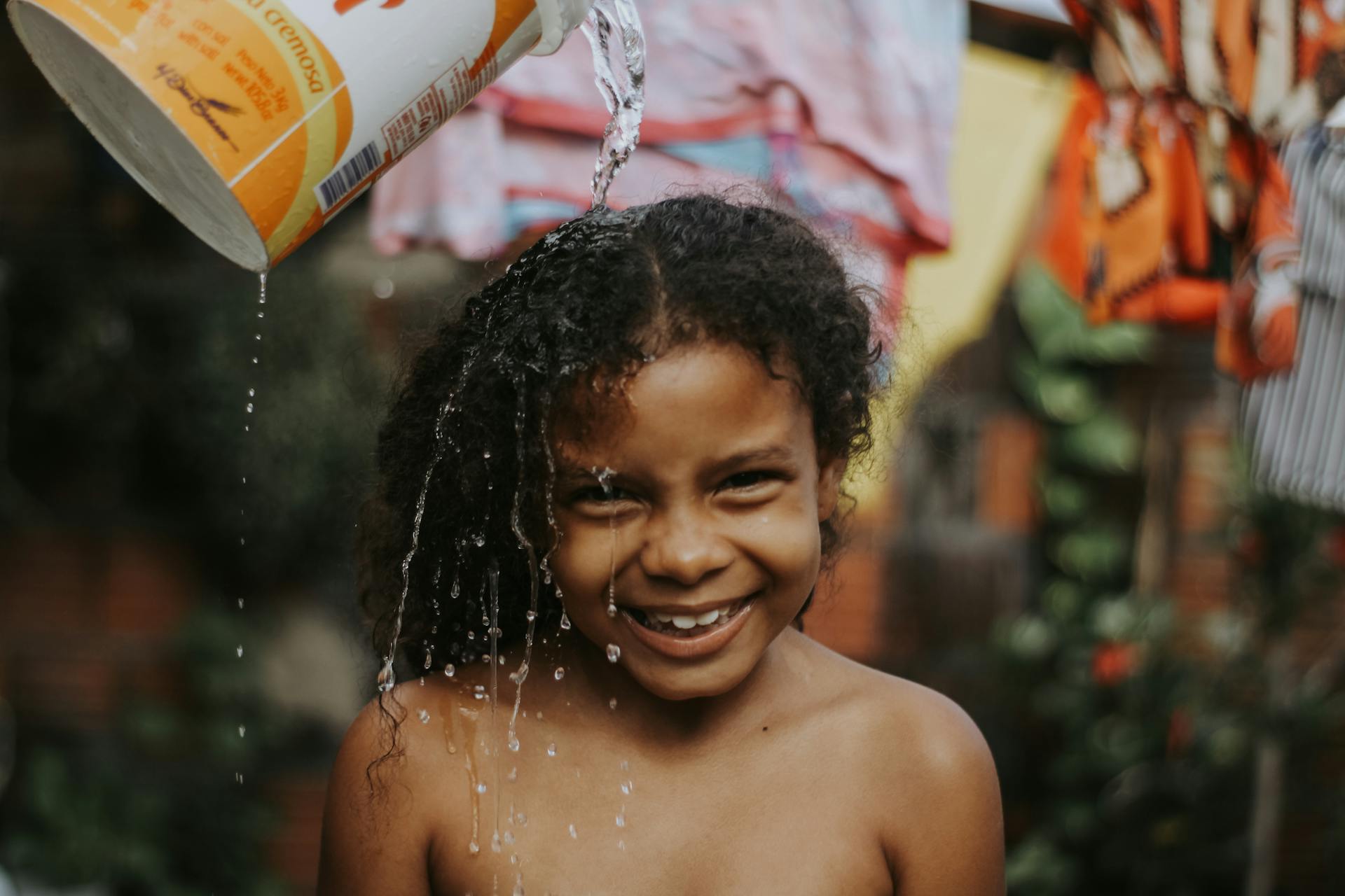 Smiling child with curly hair enjoying water being poured outdoors on a sunny day.