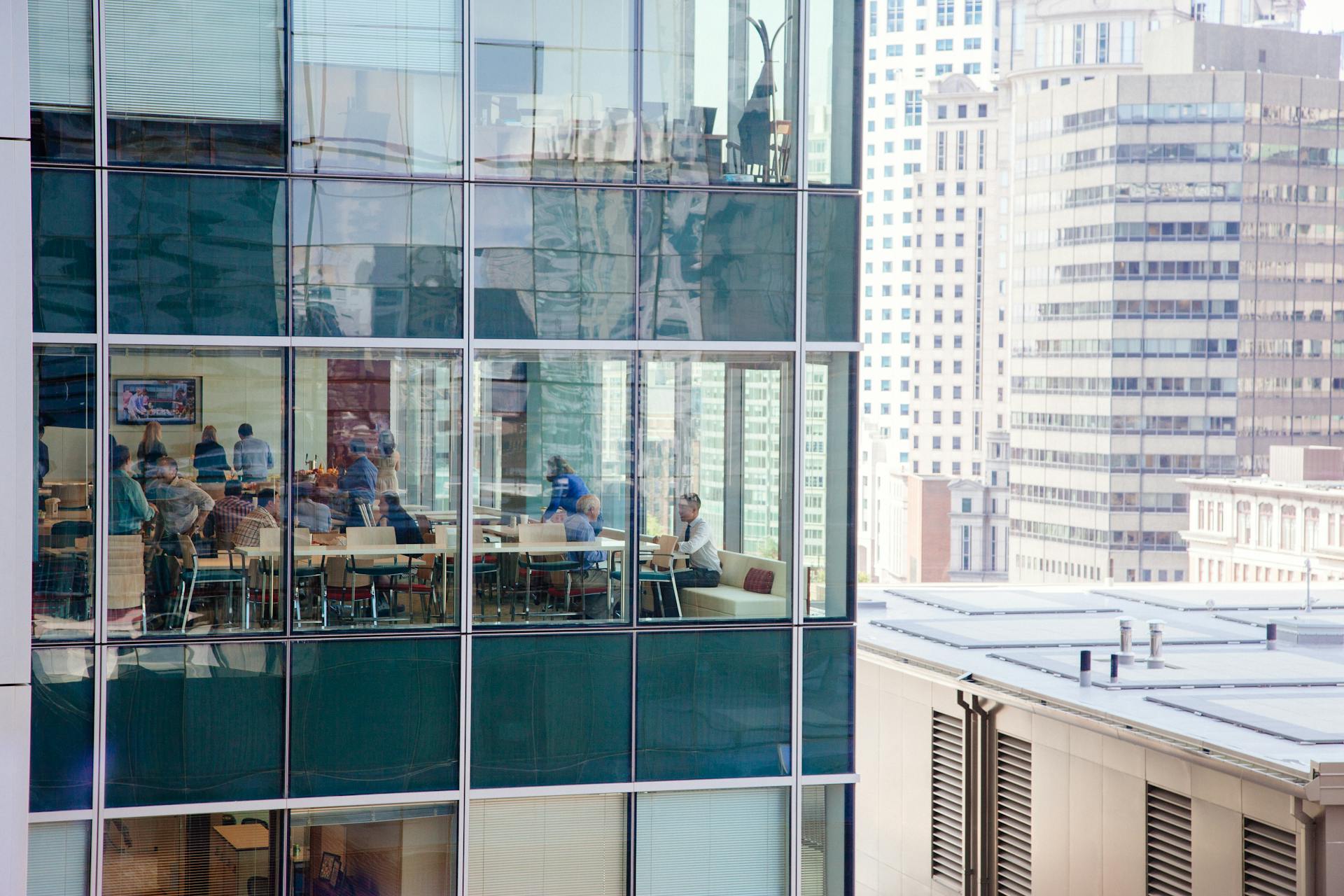 Urban office building with a glass facade reflecting New York City skyline and interior workspace activity.