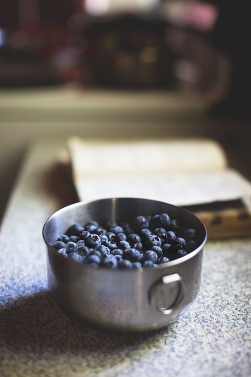 Free A metal bowl filled with blueberries placed on a kitchen counter with a cookbook in the background. Stock Photo