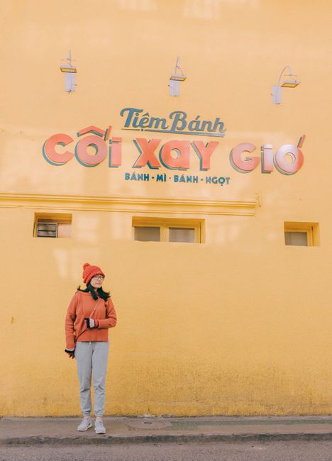 Woman stands against colorful Vietnamese bakery wall, wearing warm clothes.