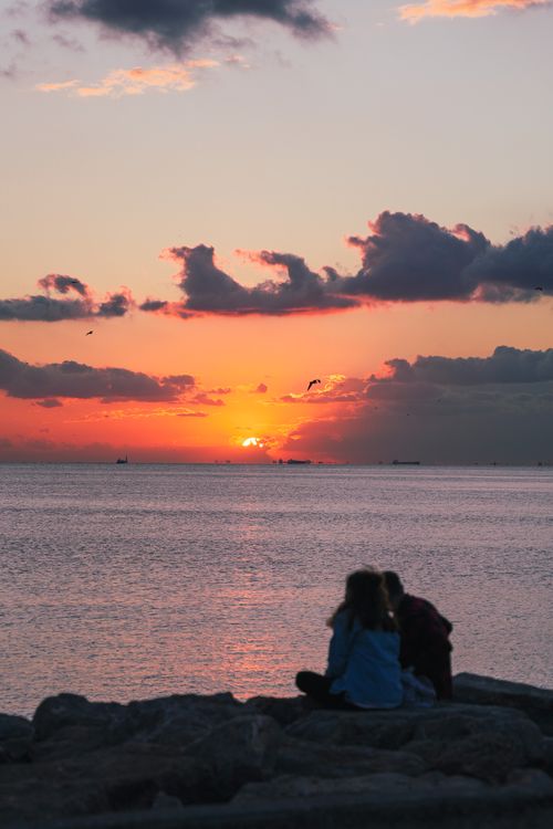 Free A couple sits on rocks by the sea, admiring a vibrant sunset with scattered clouds. Stock Photo