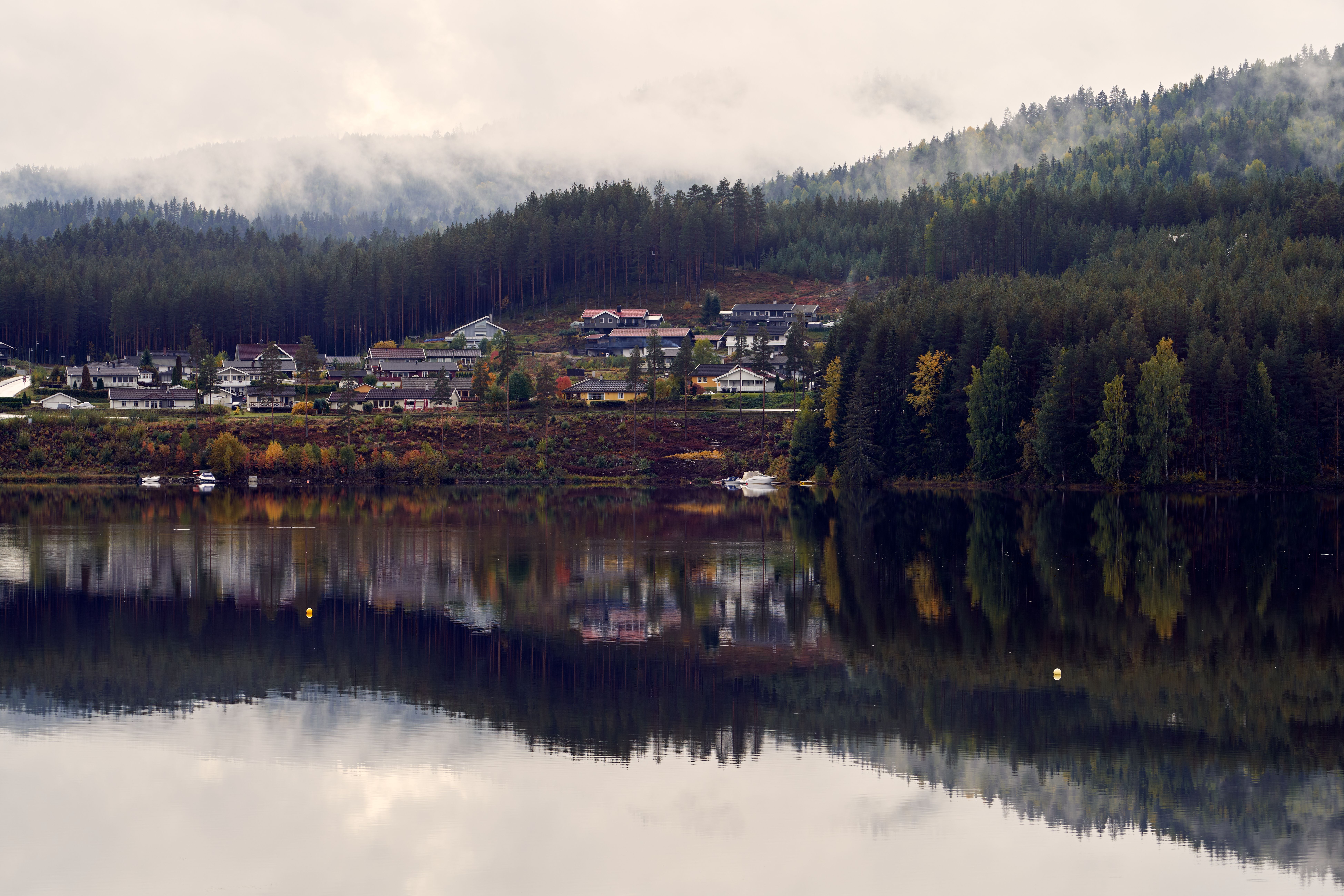 Peaceful village by a calm lake with misty mountain background and forest reflections.