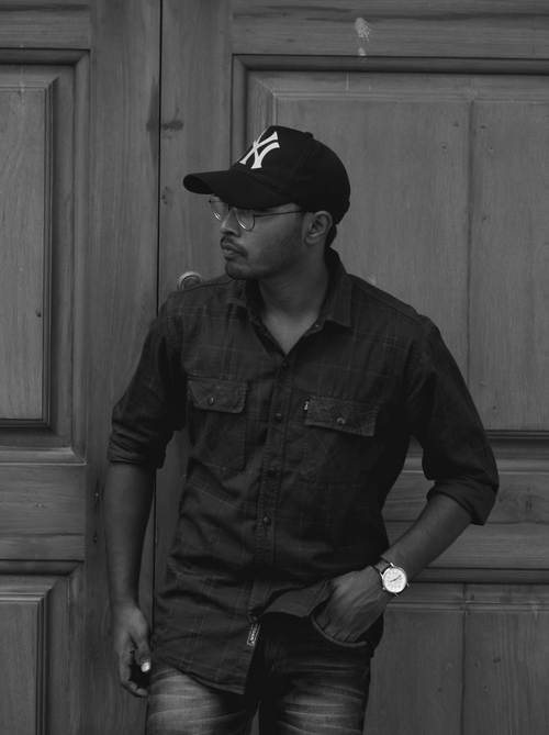 Free Man in a black shirt and cap stands against a wooden door, exuding modern fashion vibes. Stock Photo