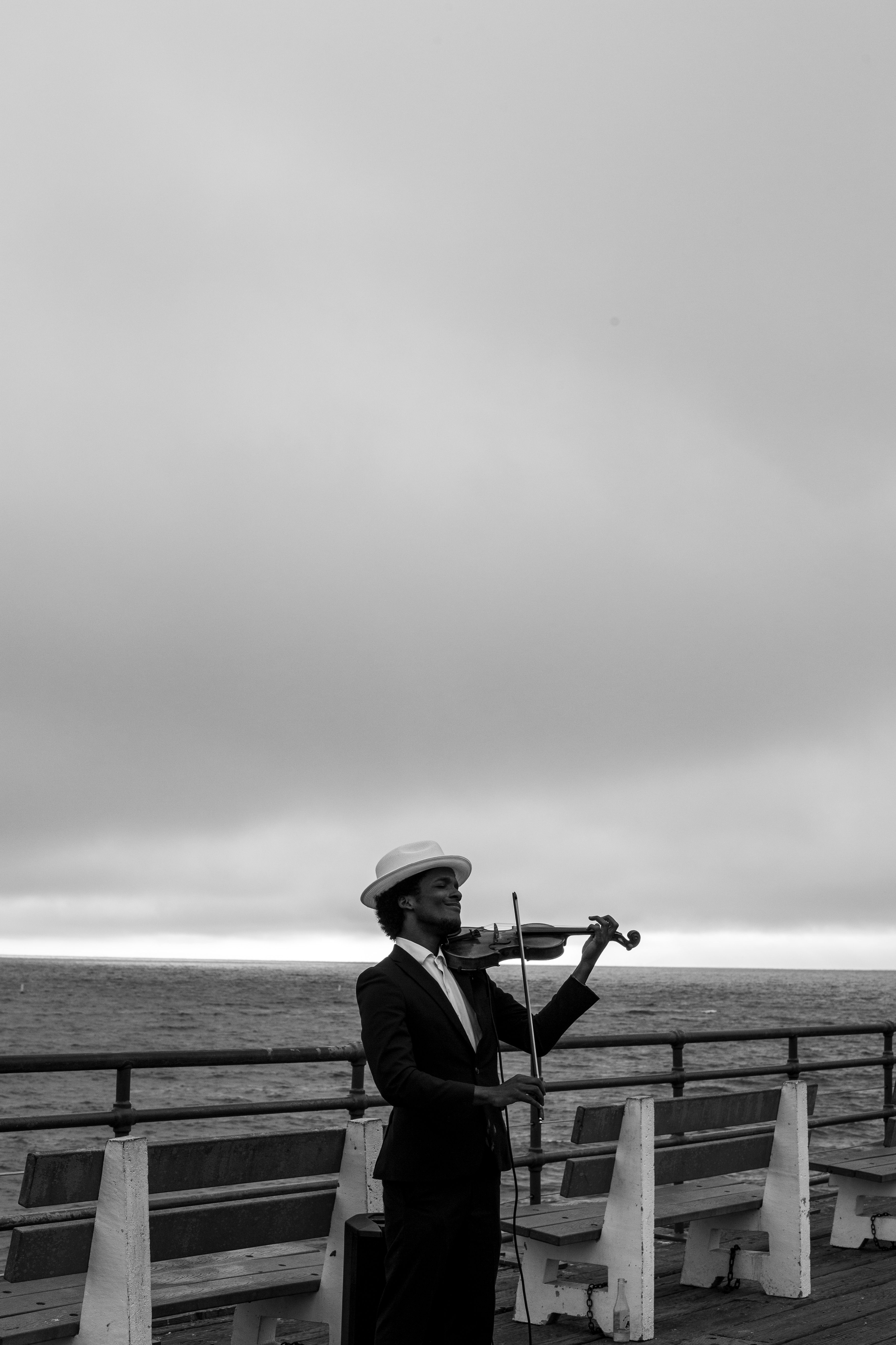 A violinist in a hat performing on a pier under cloudy skies, captured in black and white.