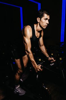 Muscular man in activewear intensely exercising on a stationary bike in a dimly lit gym.