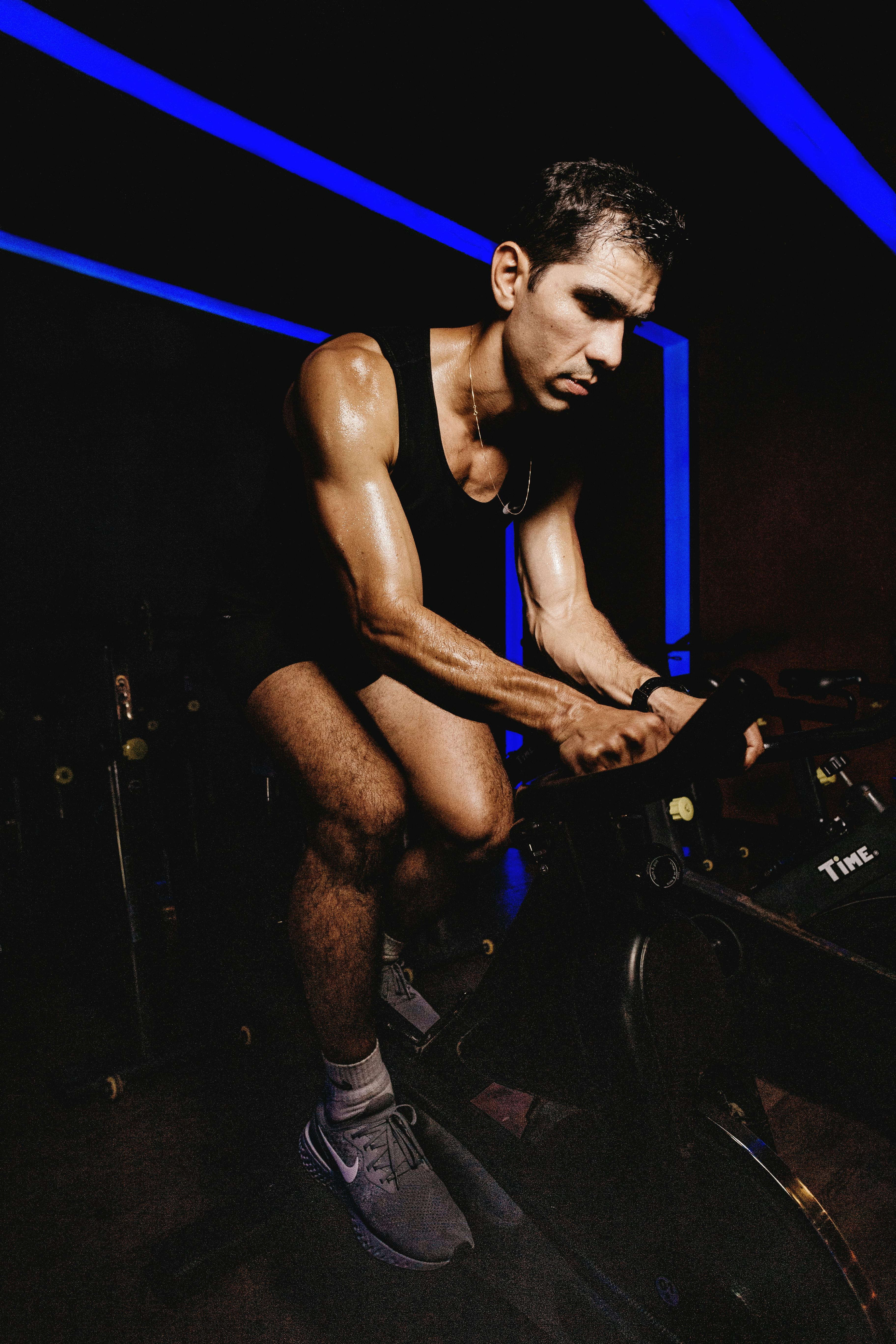 A fit man focused on an indoor cycling workout in a dimly lit gym environment.