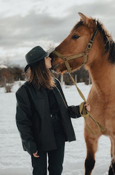 A woman wearing a hat interacts with a horse in a snowy field during winter.