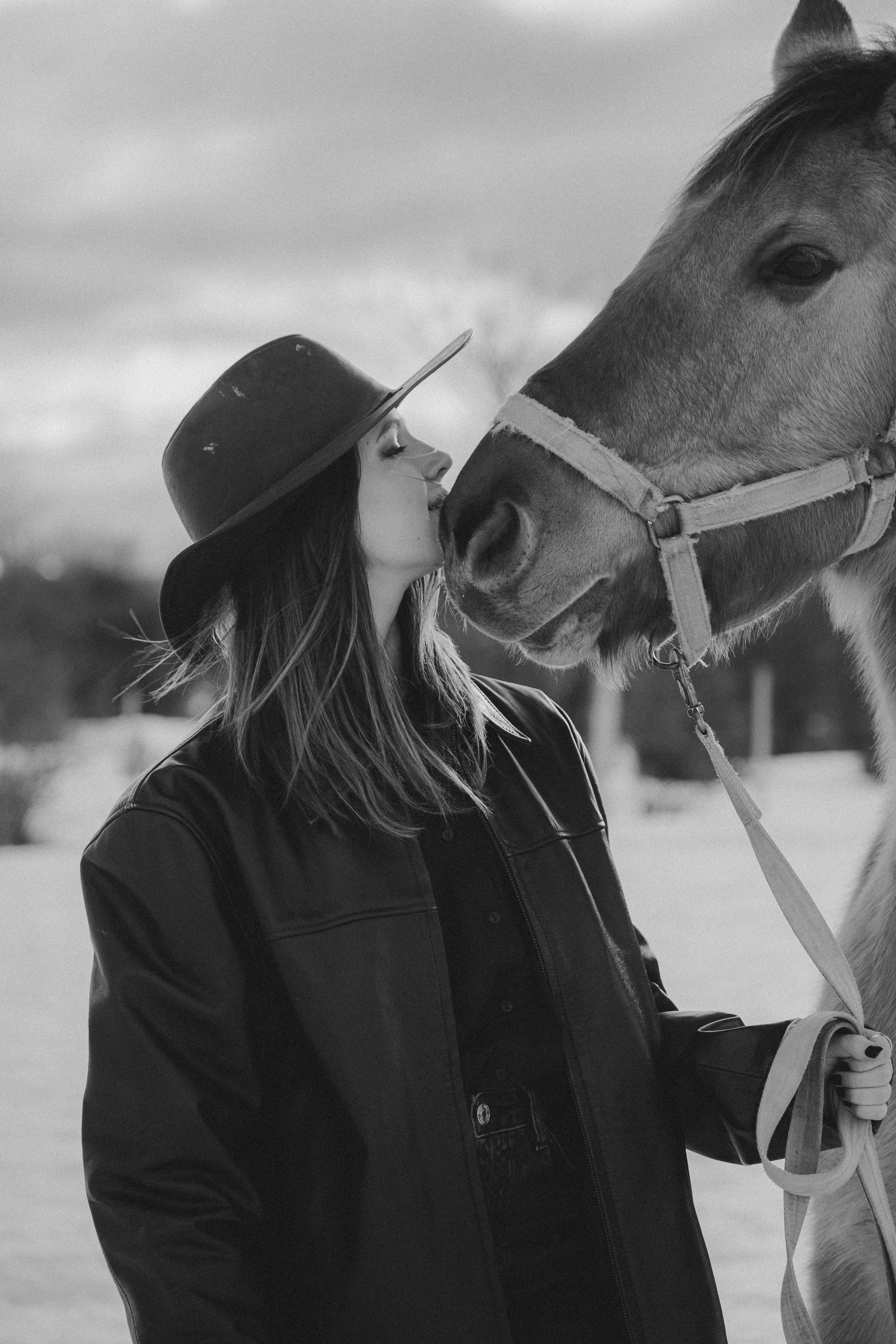 Black and white photo of a woman in cowboy hat kissing a horse outdoors in winter.