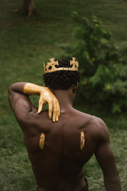 Back View of a Shirtless Man Wearing a Crown and Hands Painted with Gold Paint 