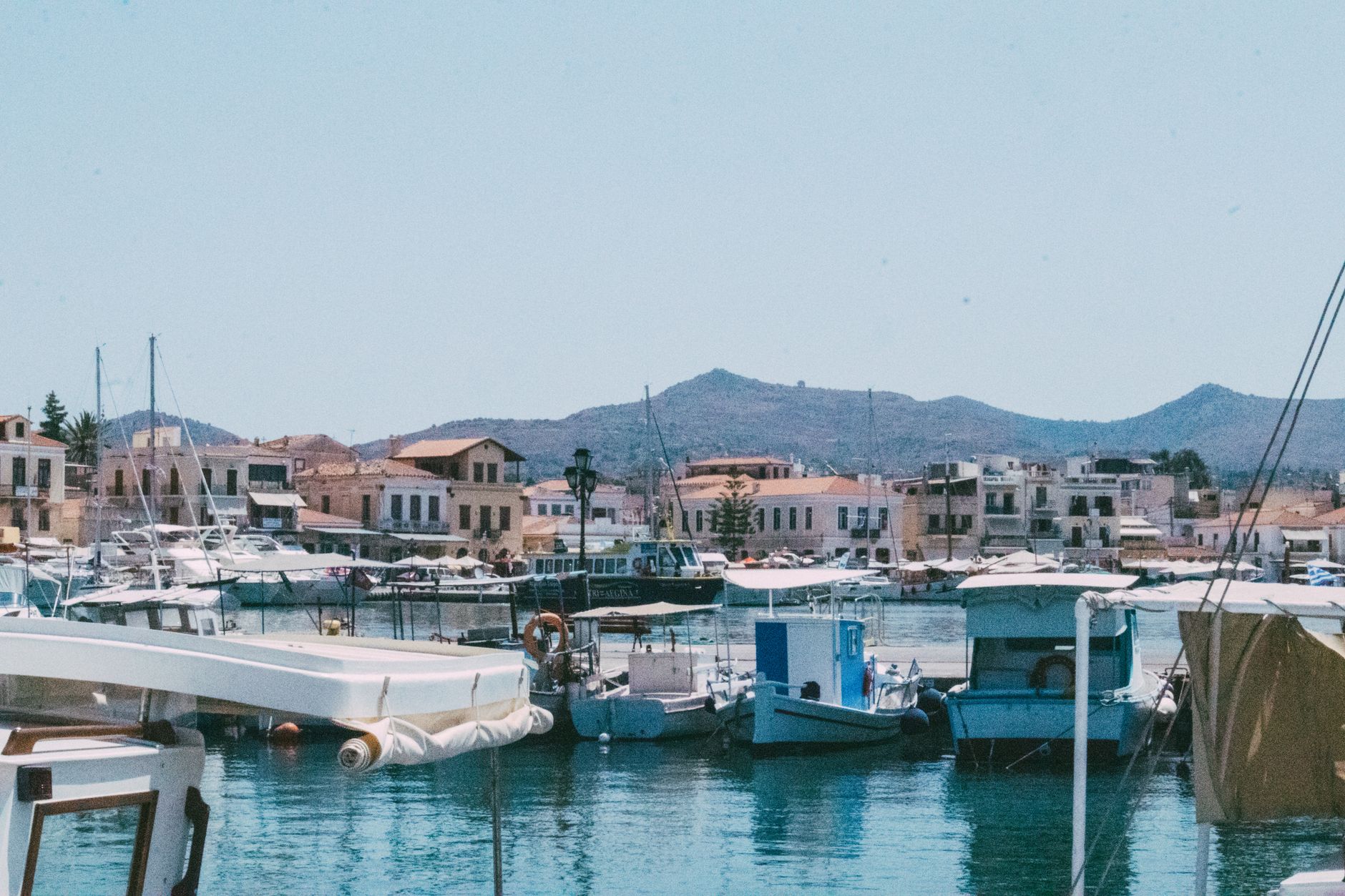 The harbour and hillside of Aegina seen across the water, with boats moored along the quay.