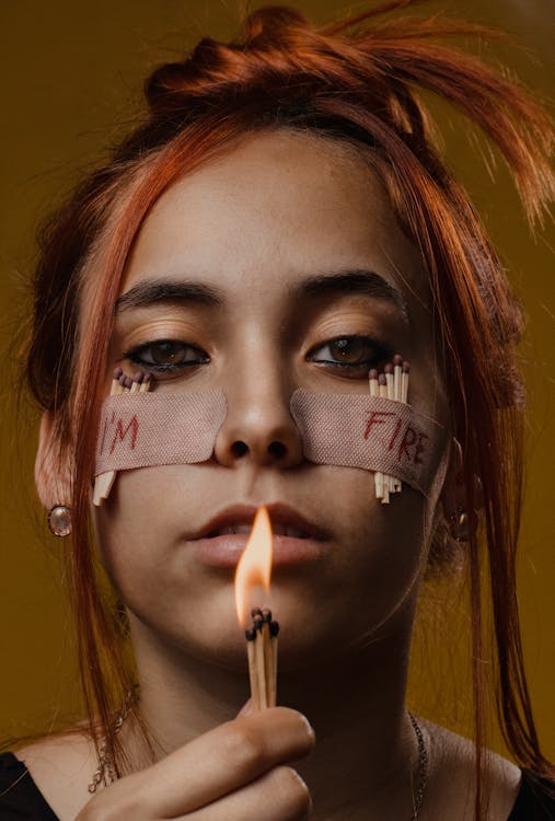 Woman with Band-aids and Matchsticks Taped to her Face Holding Burning Matchsticks