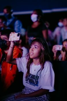 A teenager captures a live concert using her smartphone, surrounded by an audience.