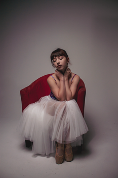 Ballet dancer wearing a white tutu, gracefully posing on a red chair in a studio.