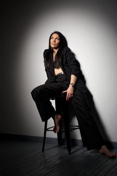 A stylish woman in black attire poses barefoot on a chair in a studio setting.