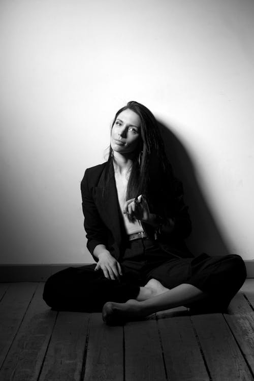 Grayscale Photo of a Beautiful Woman in Black Blazer and Black Pants Sitting on Wooden Floor while Leaning on White Wall