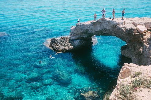 Free Tourists enjoying a sunny day cliff diving at a stunning natural rock arch over clear blue waters. Stock Photo