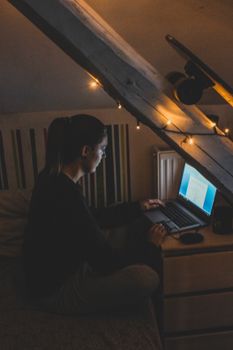 Woman working on a laptop in a dimly lit attic room with fairy lights.