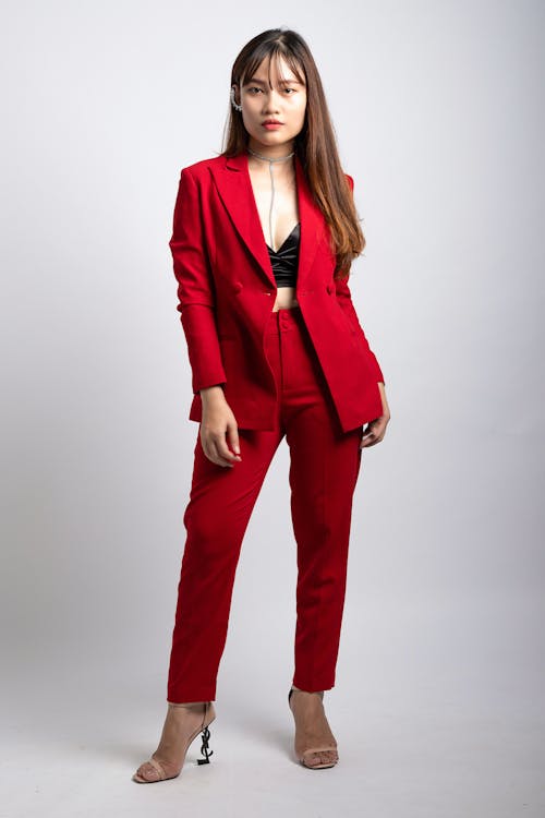 Woman Wearing Red Coat and Dress Pants Standing in Front of White Wallpaper