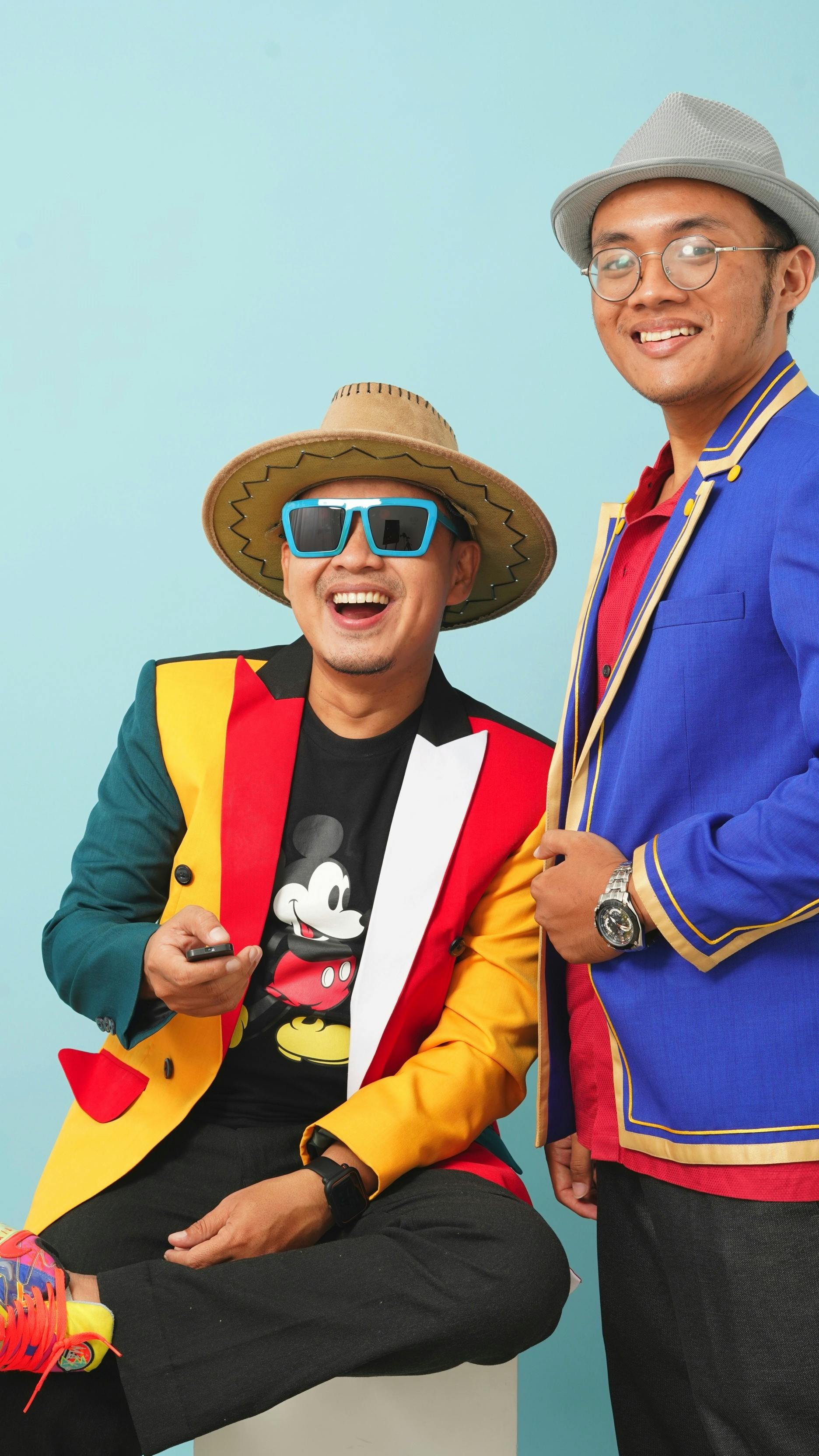 Two men laughing and posing in vibrant jackets against a blue background, exuding fun and humor.