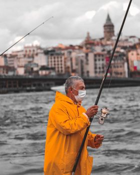 Elderly man fishing in a city wearing a mask and yellow jacket by the water.