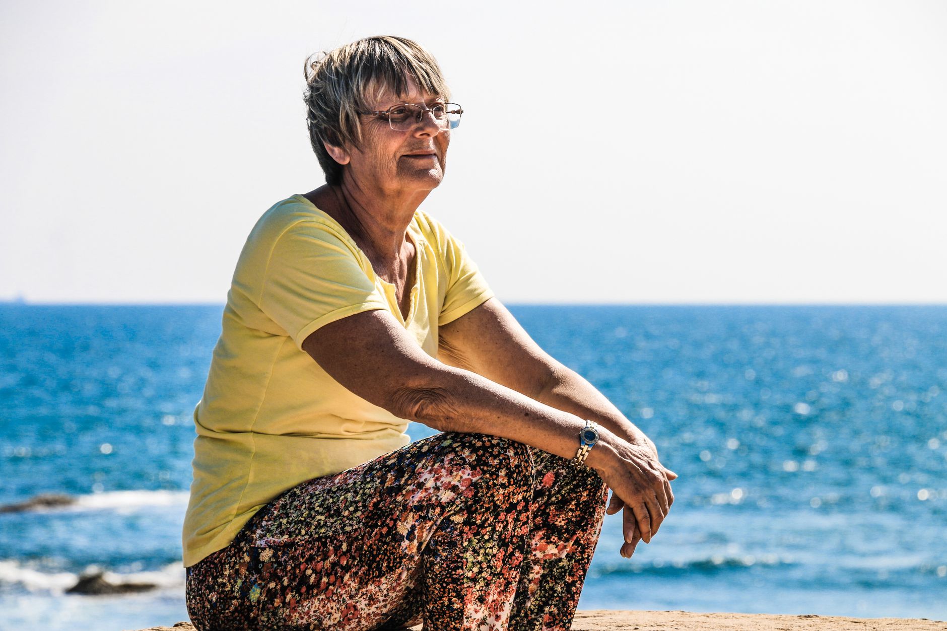 Senior woman in a yellow shirt sitting by the ocean, enjoying a sunny day.