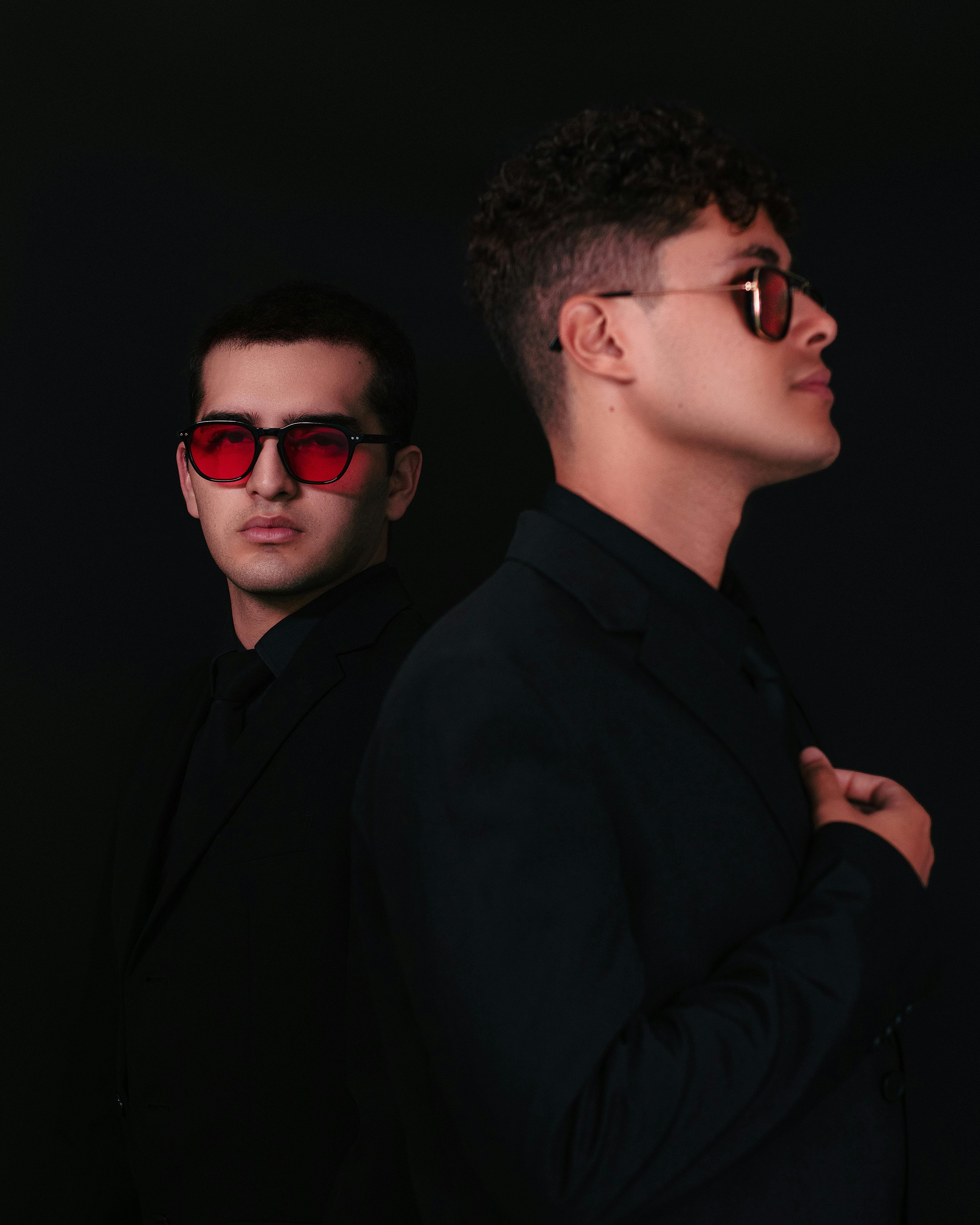 Portrait of two fashionable men in black suits and sunglasses against a black background.