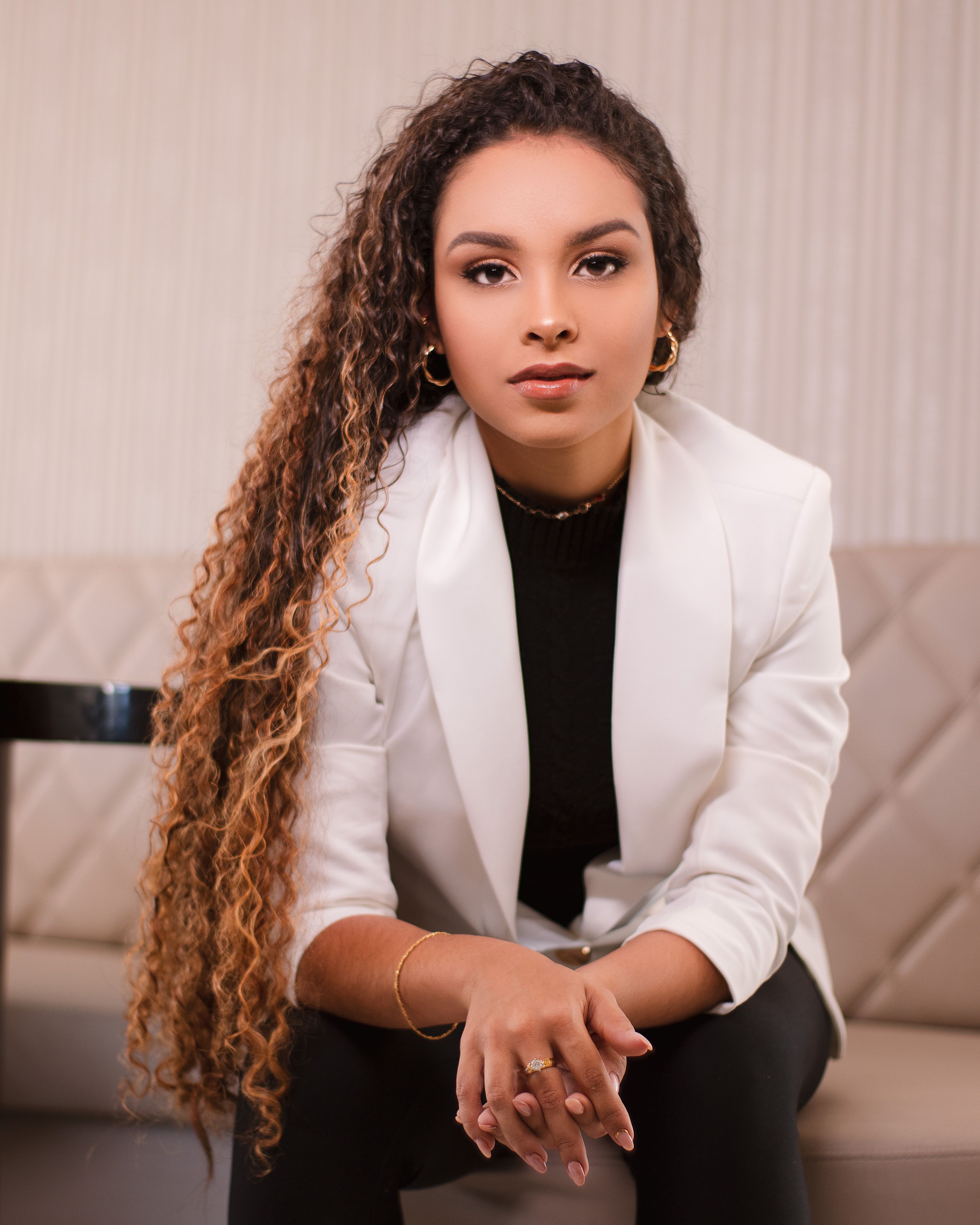 Portrait of a confident woman with curly hair sitting indoors in a white blazer.