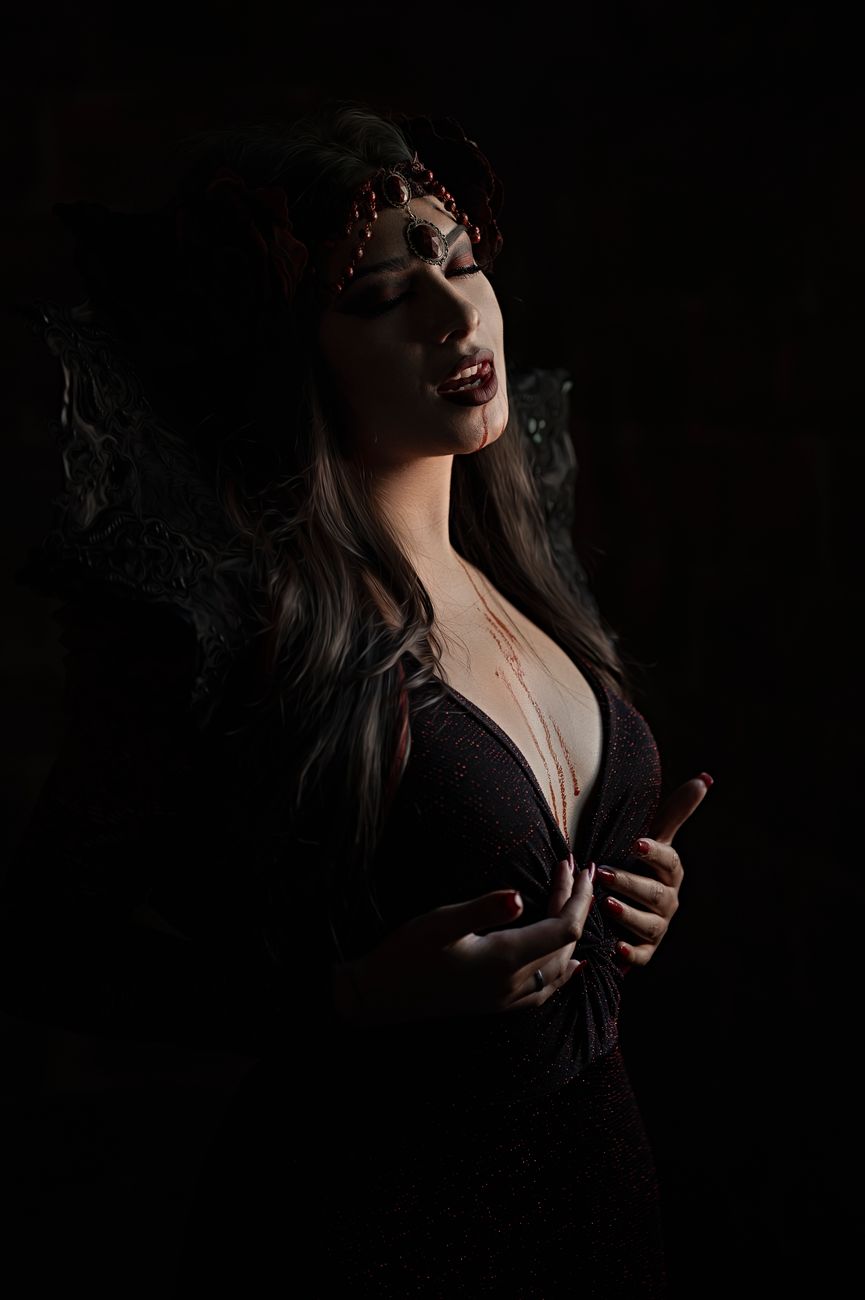 A woman in dark attire, adorned in jewels, captivates with a mysterious pose in deep shadows.