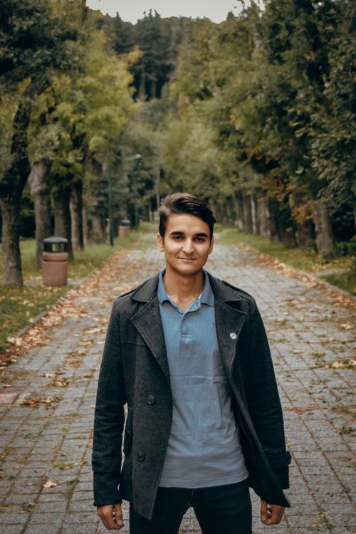 Free Young man wearing a gray blazer, smiling on a tree-lined pathway outdoors. Stock Photo