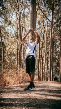 A young man wearing a white shirt stands on a sunny forest trail, looking upwards.