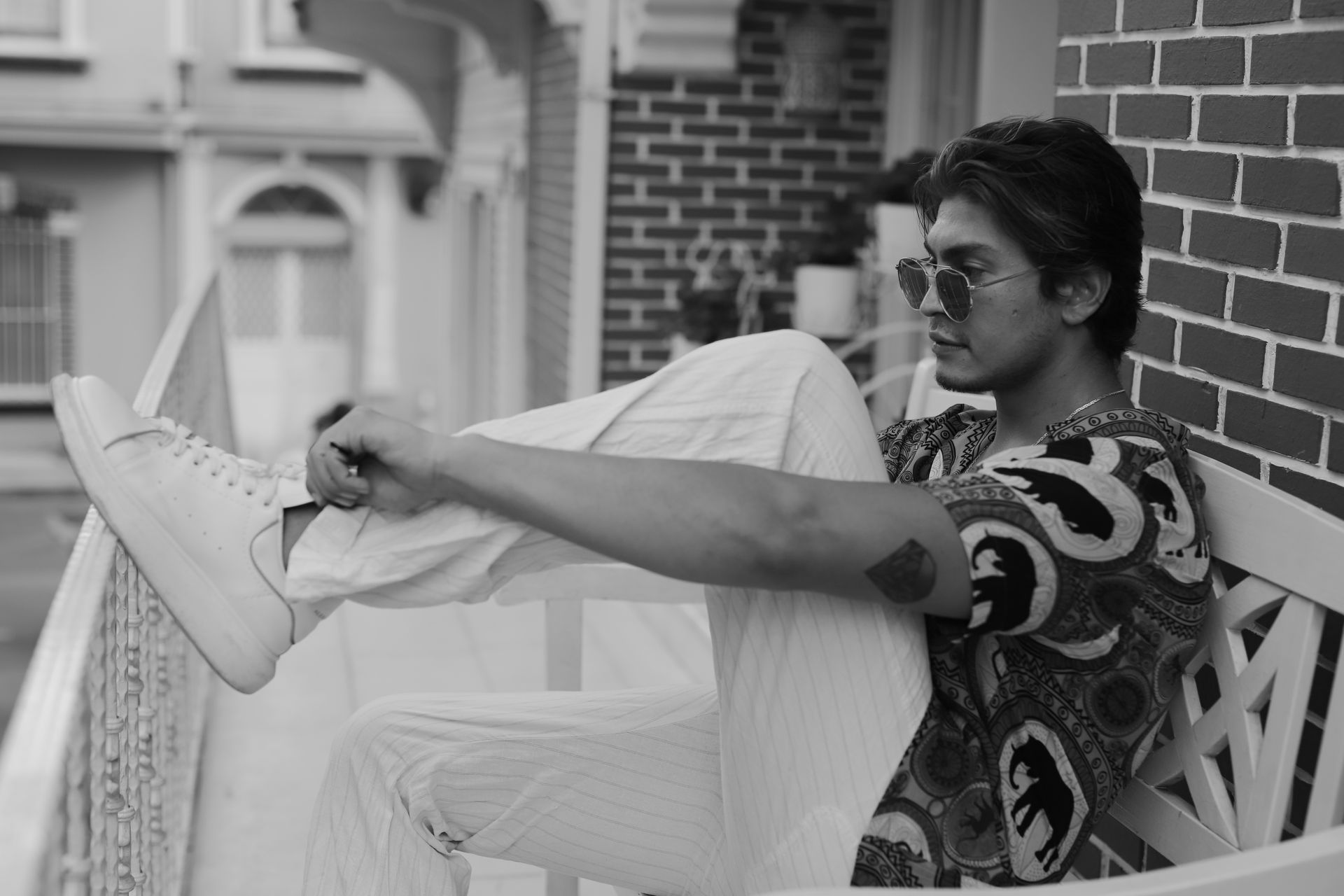 A fashionable man wearing sunglasses and striped pants adjusts his sneakers while sitting on a balcony.