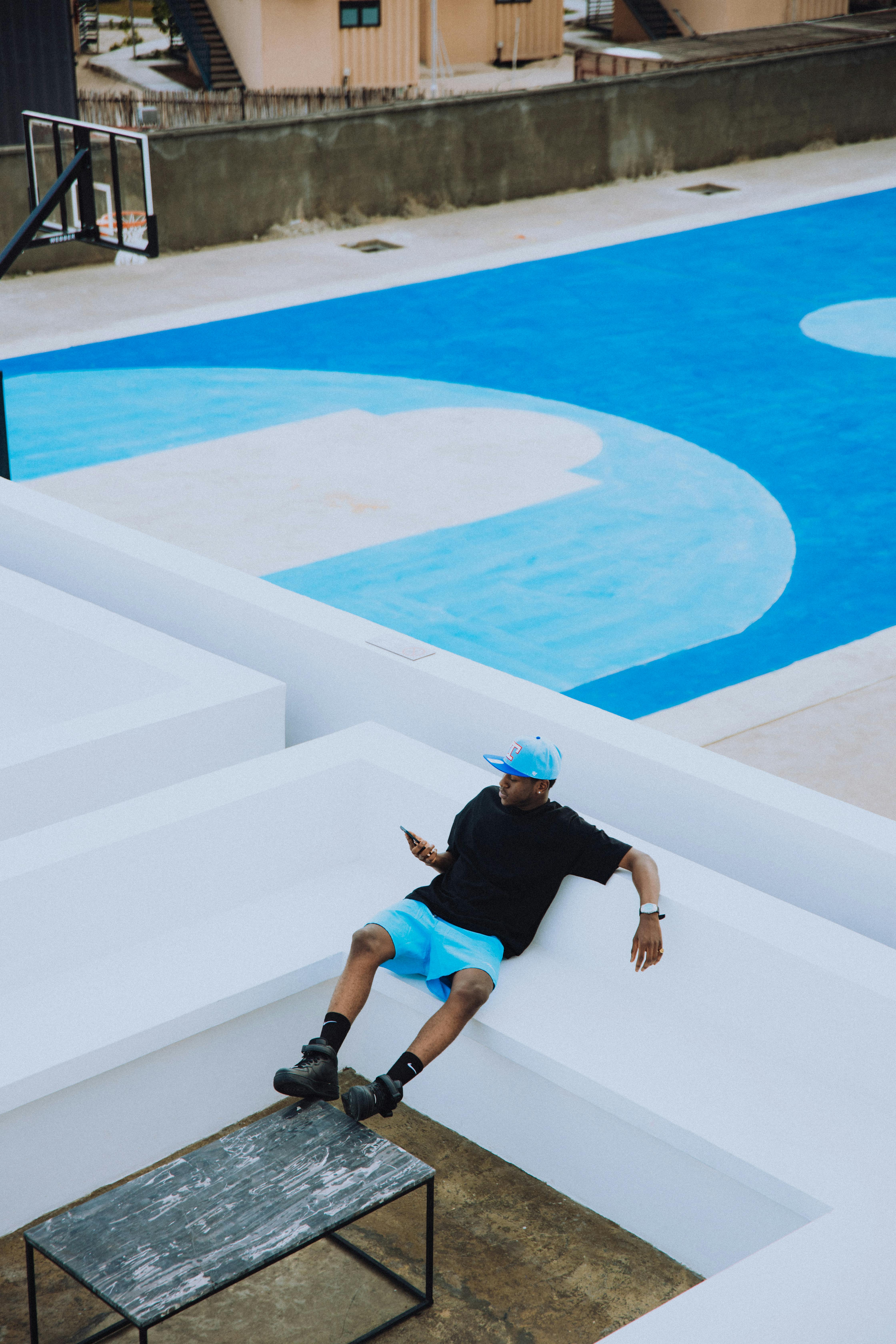 A man in casual attire sits on a white geometric bench near a vibrant blue basketball court.