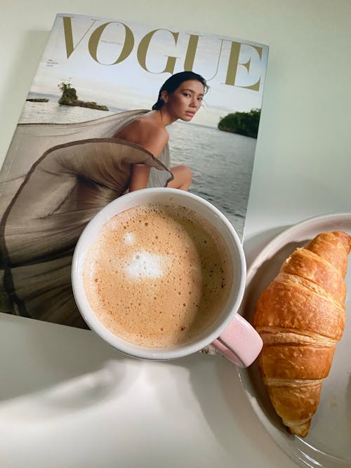 Croissant on a Plate Beside Cup of Coffee and Magazine