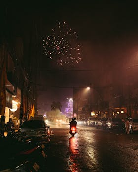 A bustling urban street illuminated by fireworks lighting up the night sky. Vibrant and lively atmosphere.