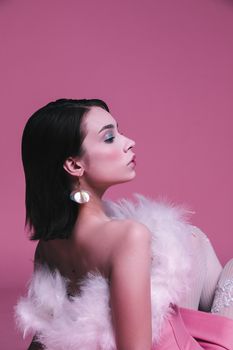 A woman in a pink dress and feathered top poses against a pink background, creating an elegant and dreamy mood.