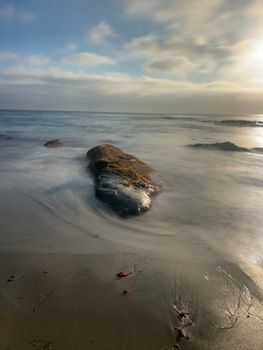A peaceful coastal scene with rocks, ocean waves, and a cloudy sky at sunset.