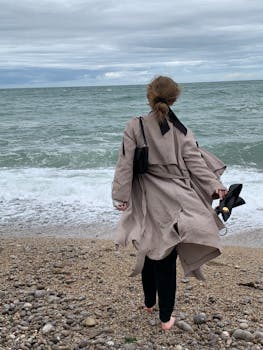 Woman in trench coat walks along a windy pebble beach holding heels, gazing at sea.