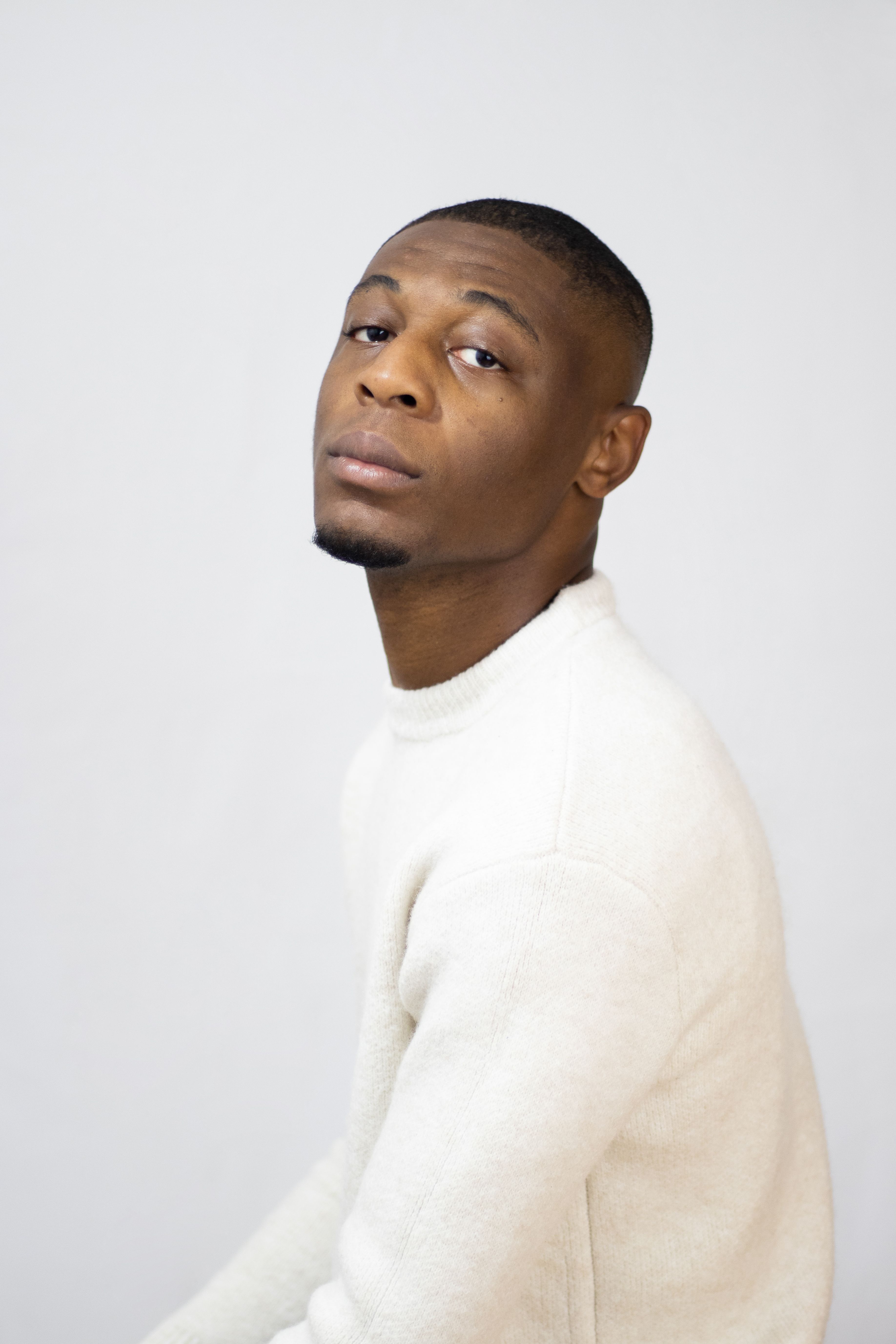 A portrait of a stylish, serious-looking man wearing a white sweater against a plain background.