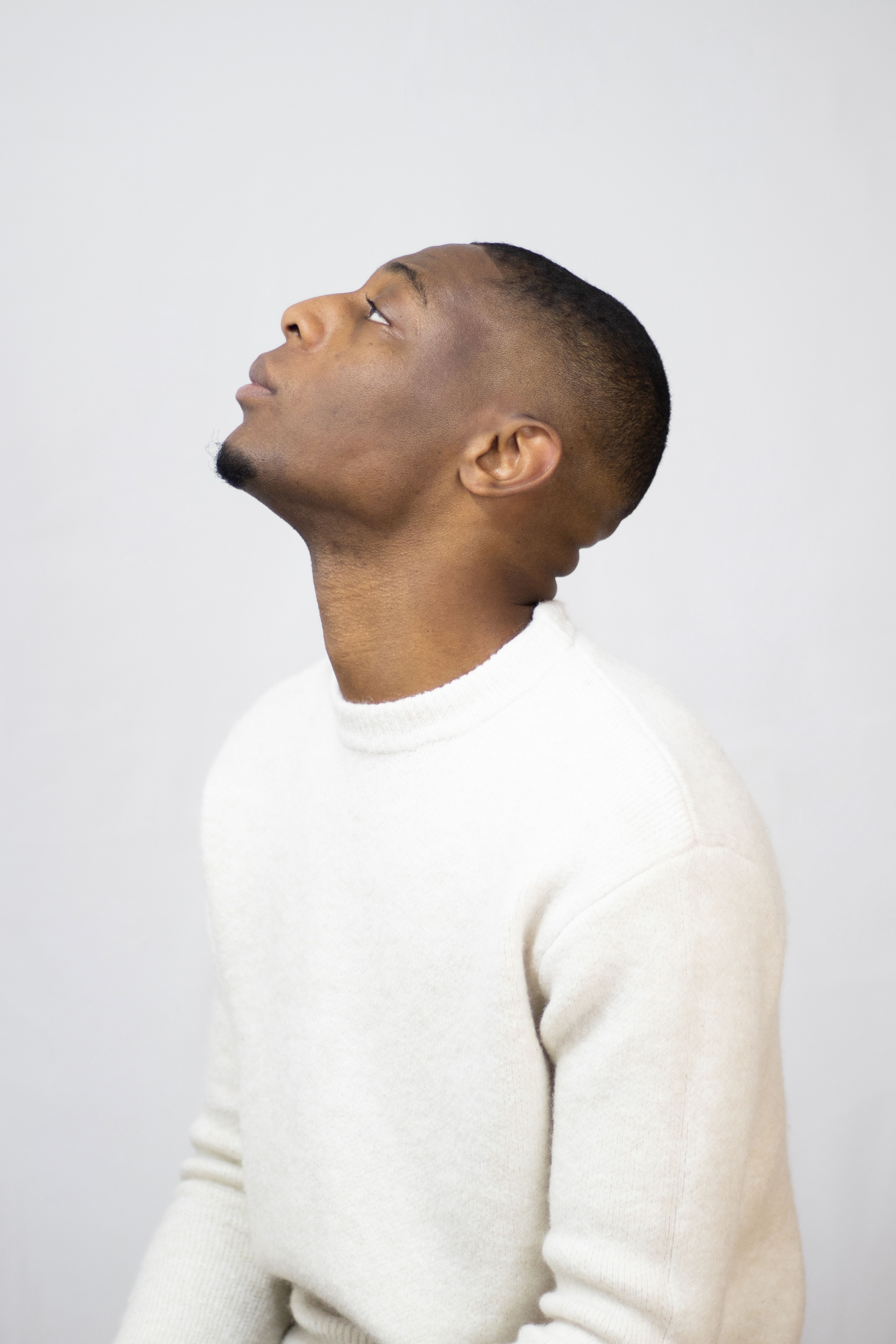 Side profile of a black man looking upwards, wearing a white sweater against a plain background.