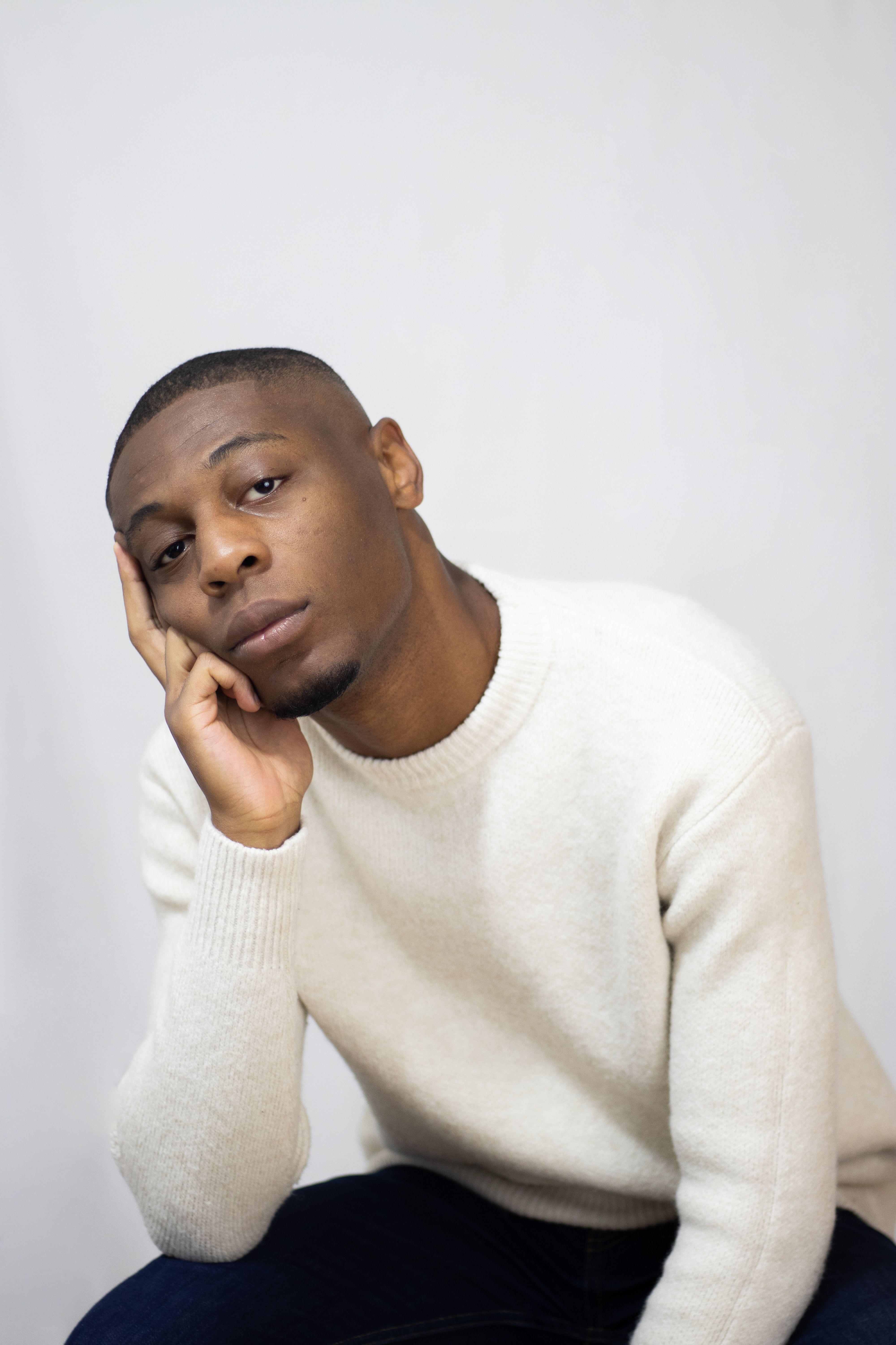 Close-up portrait of a man sitting with hand on face wearing a white sweater against a white background.