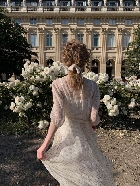 Back view of a blonde woman in a white dress walking through a rose garden.