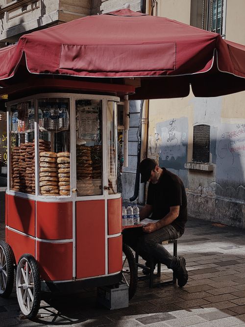 Free Mature street vendor reading beside a vibrant red food cart under an umbrella. Stock Photo