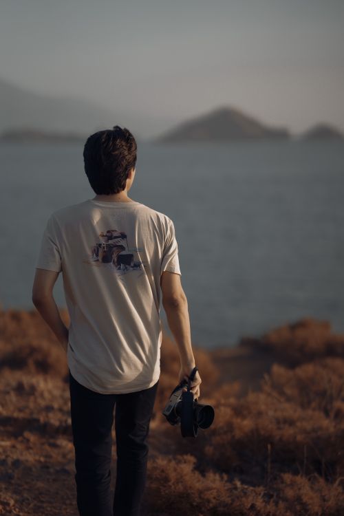 Free A man holding a camera walks on a path overlooking a calm seascape, with distant islands in view. Stock Photo
