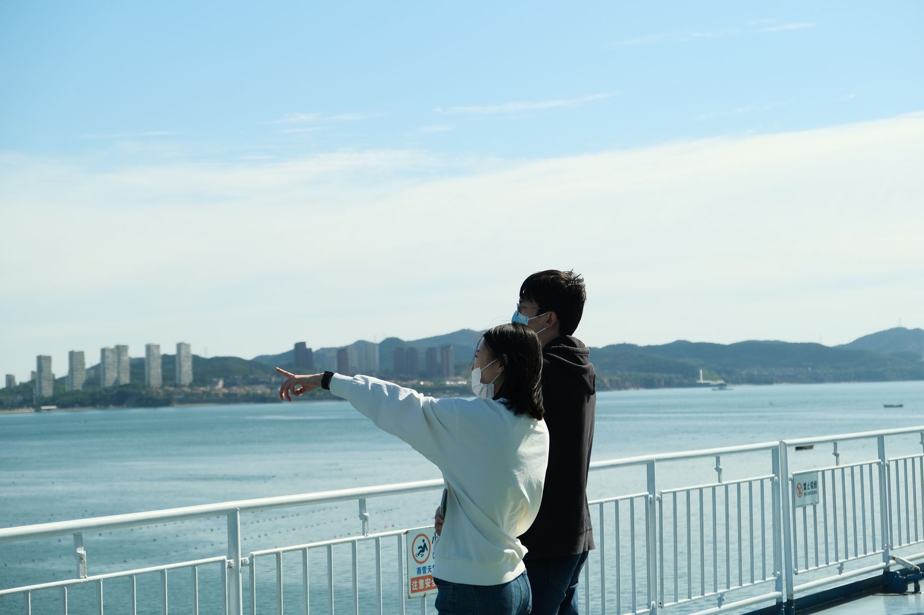 Couple wearing face masks enjoying a scenic seaside view, pointing towards the horizon.