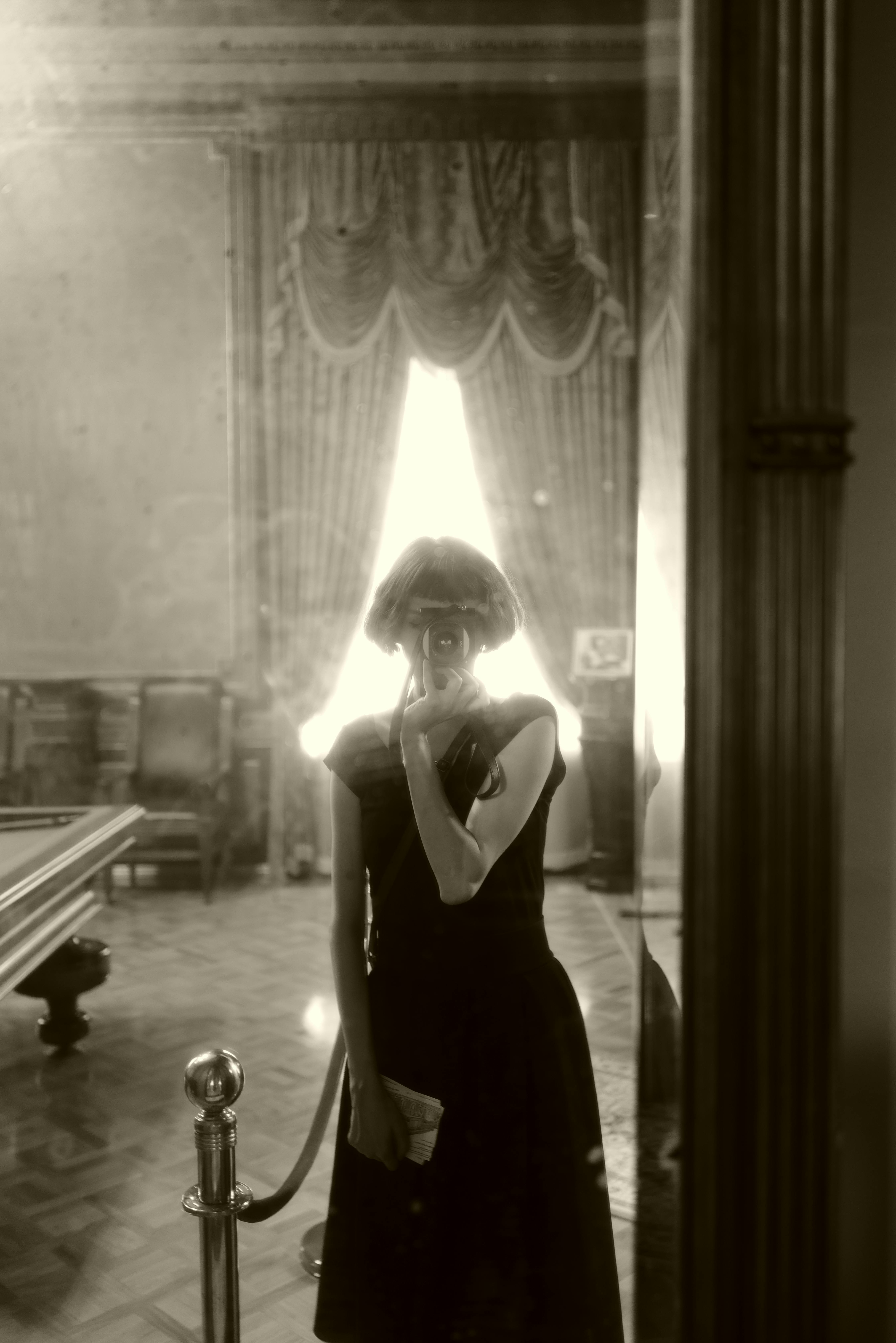A woman in a vintage dress stands in an elegant, classic interior, reflecting through a window.